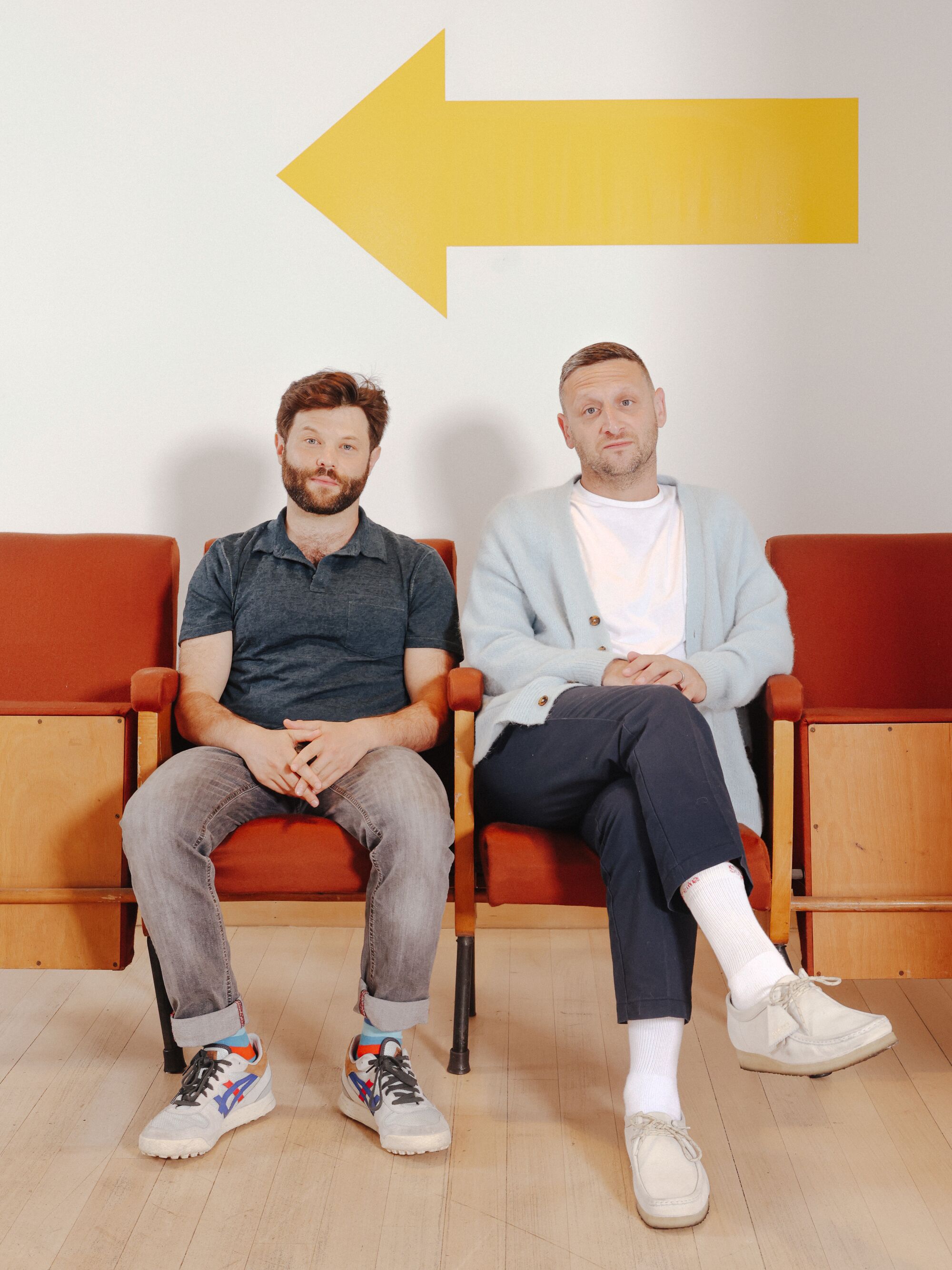 Two men sit in chairs with a large yellow arrow on the wall behind them.