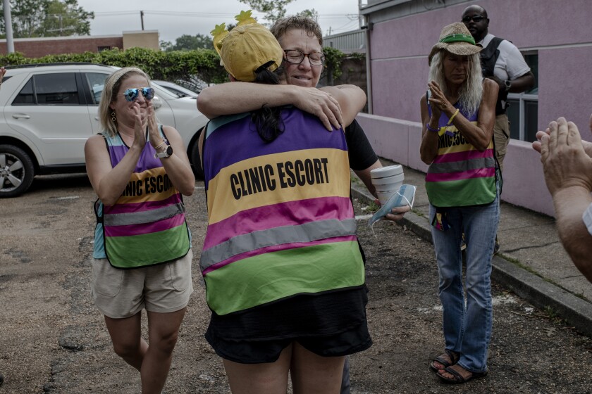 A woman wearing a colorful striped vest with the words Clinic Escort embraces another person as others clap around them