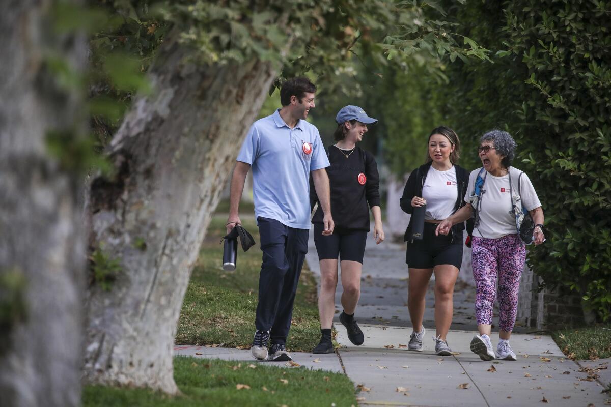 A man carrying a water bottle and three women walk along a neighborhood sidewalk alongside mature trees.