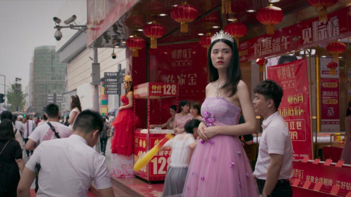 A young woman modeling a pink gown and tiara stares into the distance as people pass on a busy street. 