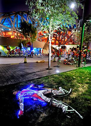 Another performer, illuminated and wearing stilts, rests outside the stadium.