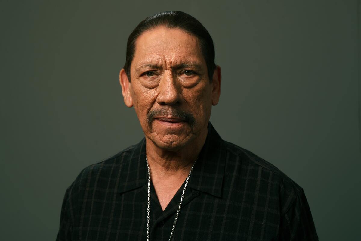 Danny Trejo looks forward, wearing a black V-neck shirt and a thin chain necklace