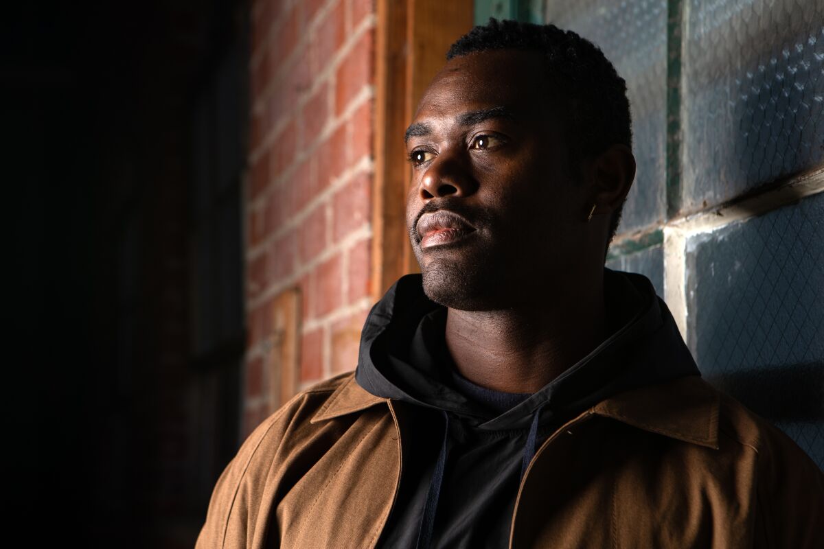 Portrait of a man standing in front of a brick wall, looking out of frame to his right. 