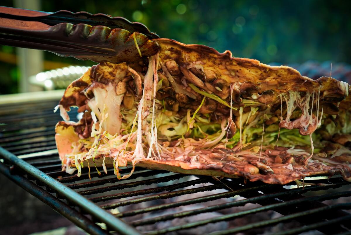 Tongs lift the top of a vegetarian tlayuda, on the grill, to reveal cheese and chopped nopales within.