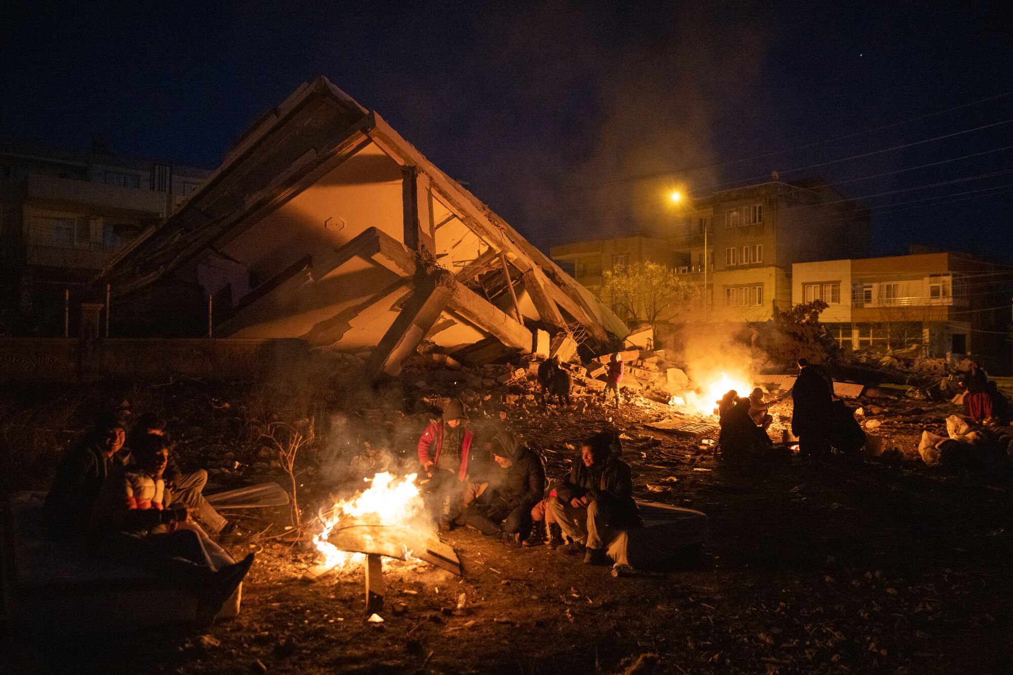 Displaced people sit around camp fires surrounded by destroyed buildings in the dark