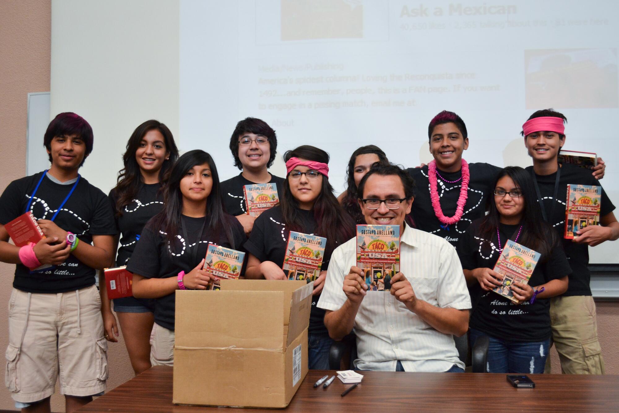 A man in glasses and a white shirt holds up a book, surrounded by people in dark shirts, some holding the same books 