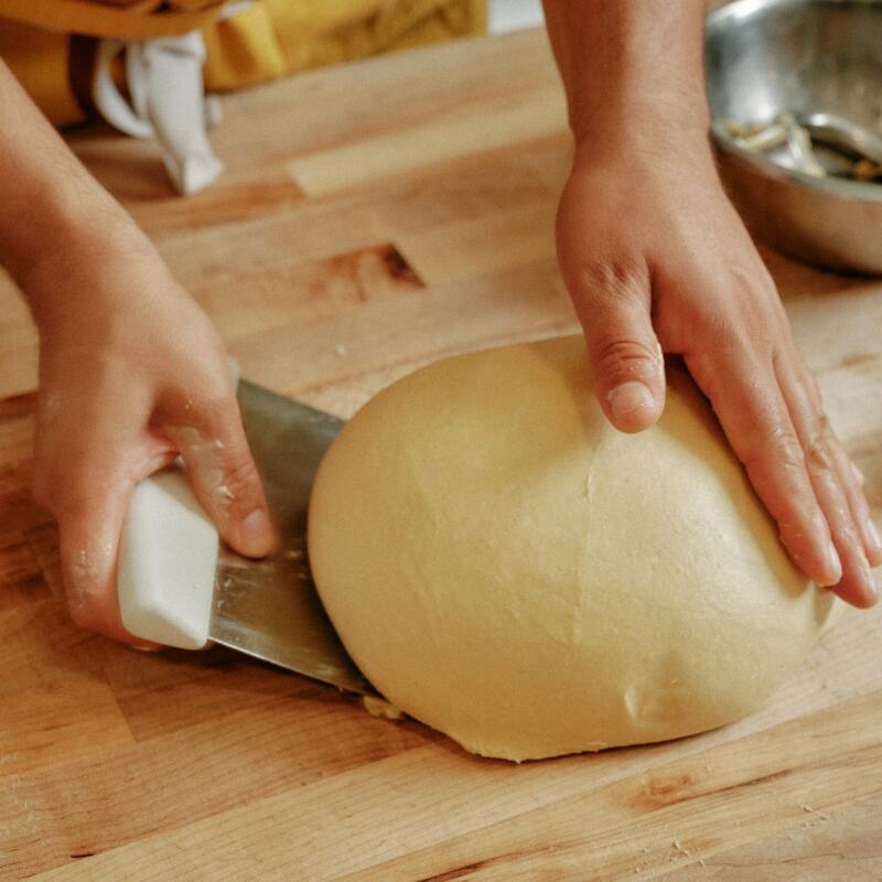 Hands use a bench scraper to pick up a smooth ball of dough