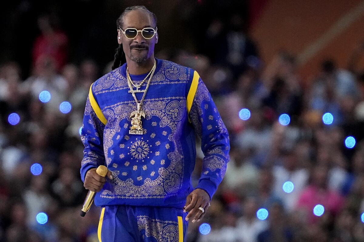 Snoop Dogg is wearing sunglasses and a blue and yellow sweater and pants as he holds a microphone by his waist