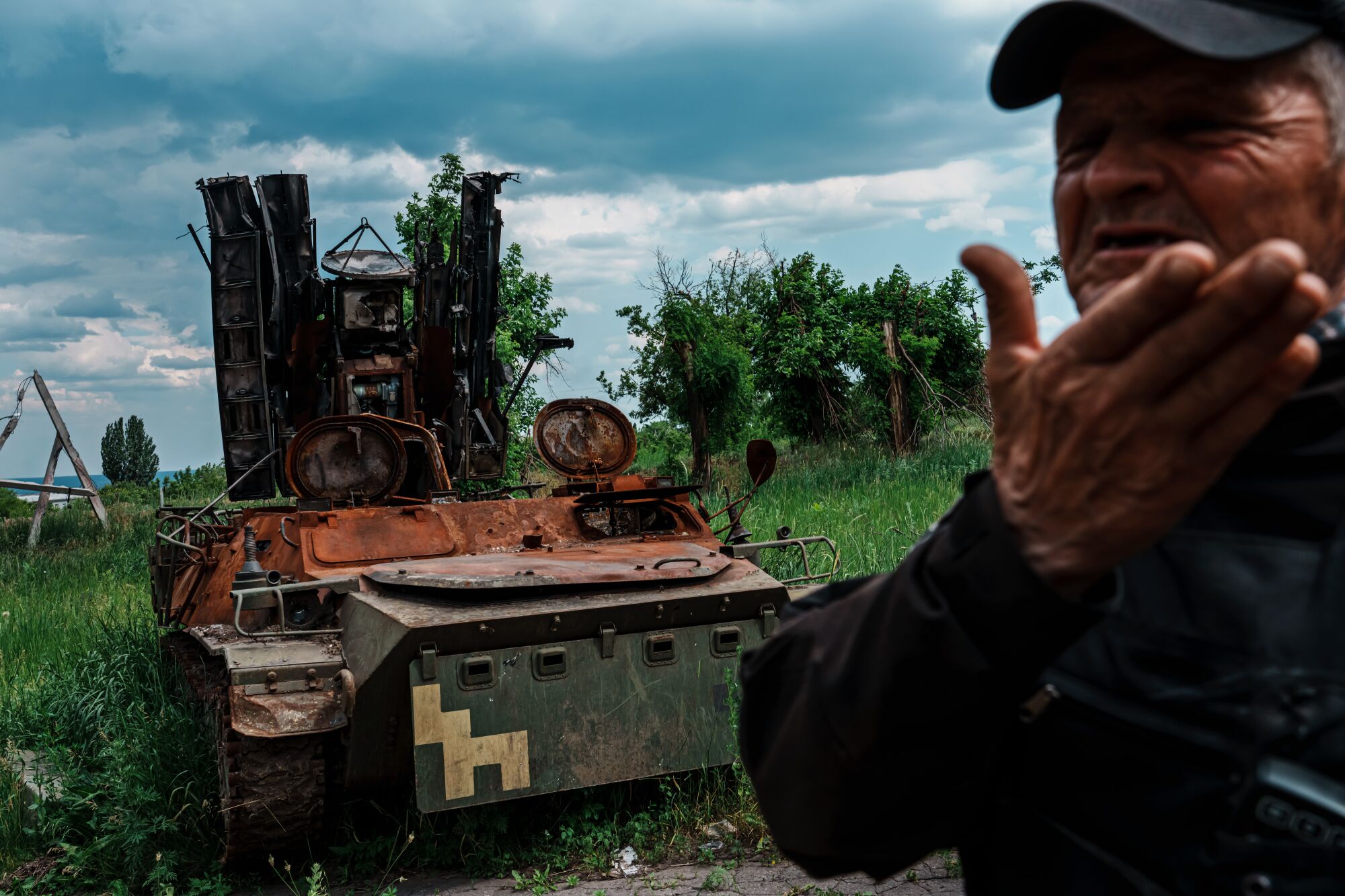 A man gestures near the hulking wreckage of a military vehicle in a grassy area 