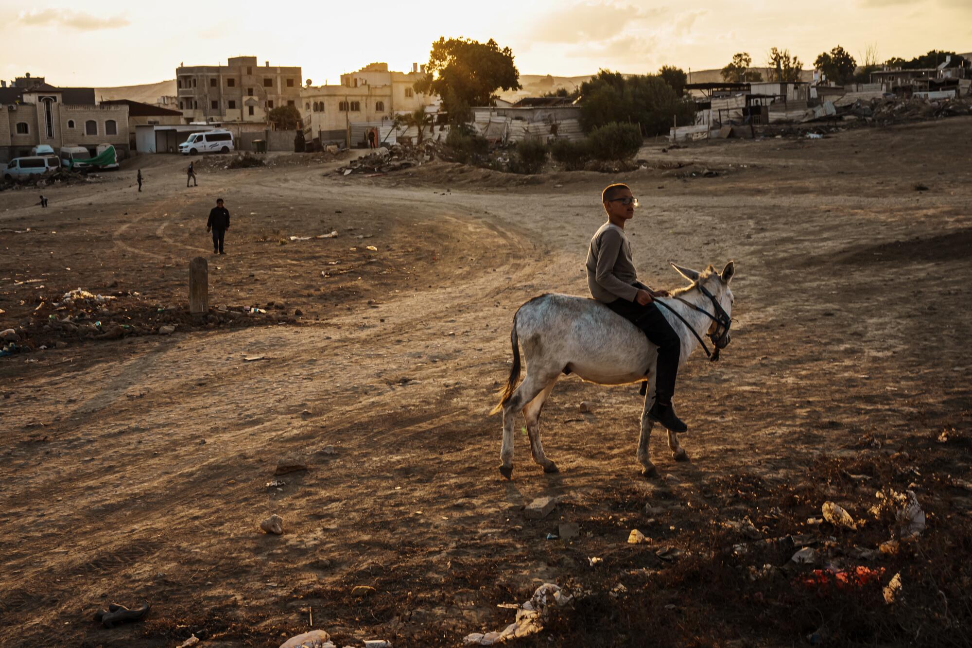 A boy rides a donkey in the desert, with a group of buildings in the background