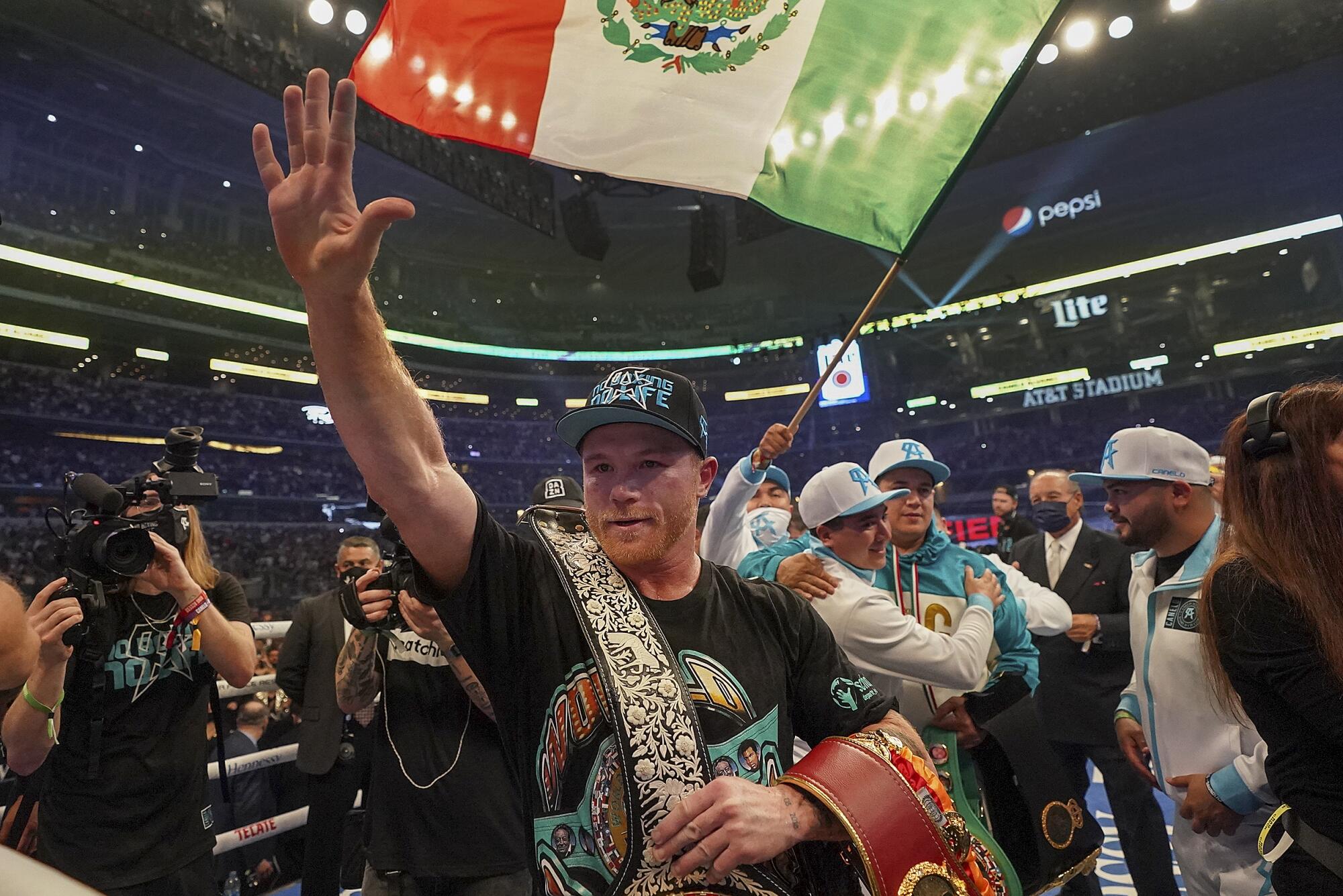 Canelo Álvarez carries his title belts in the ring as others take photos and mill around smiling