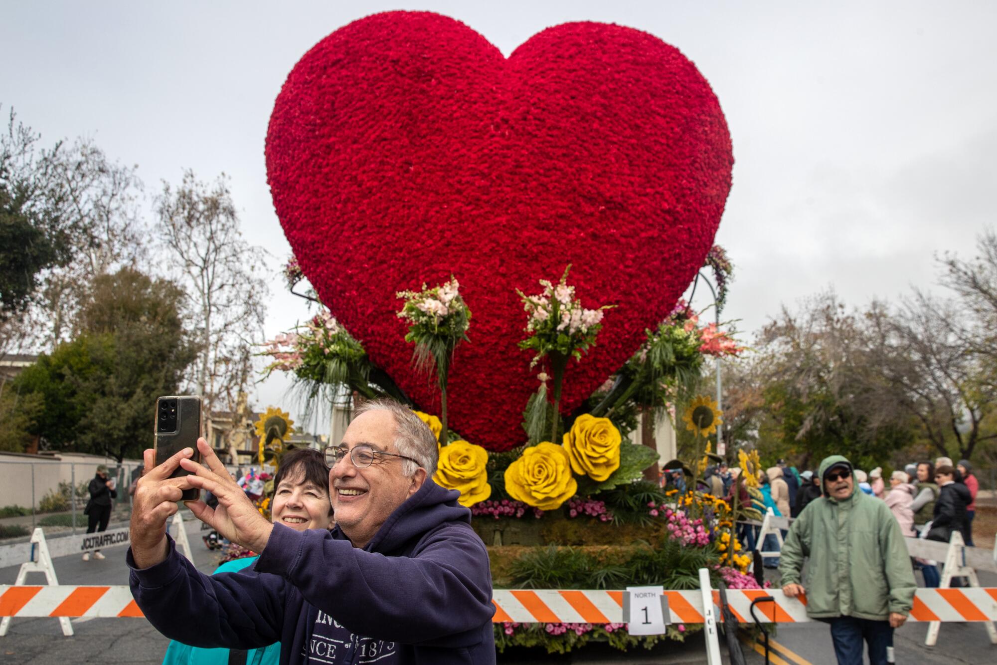 A man and a woman take a selfie in front of a heart-shaped float 