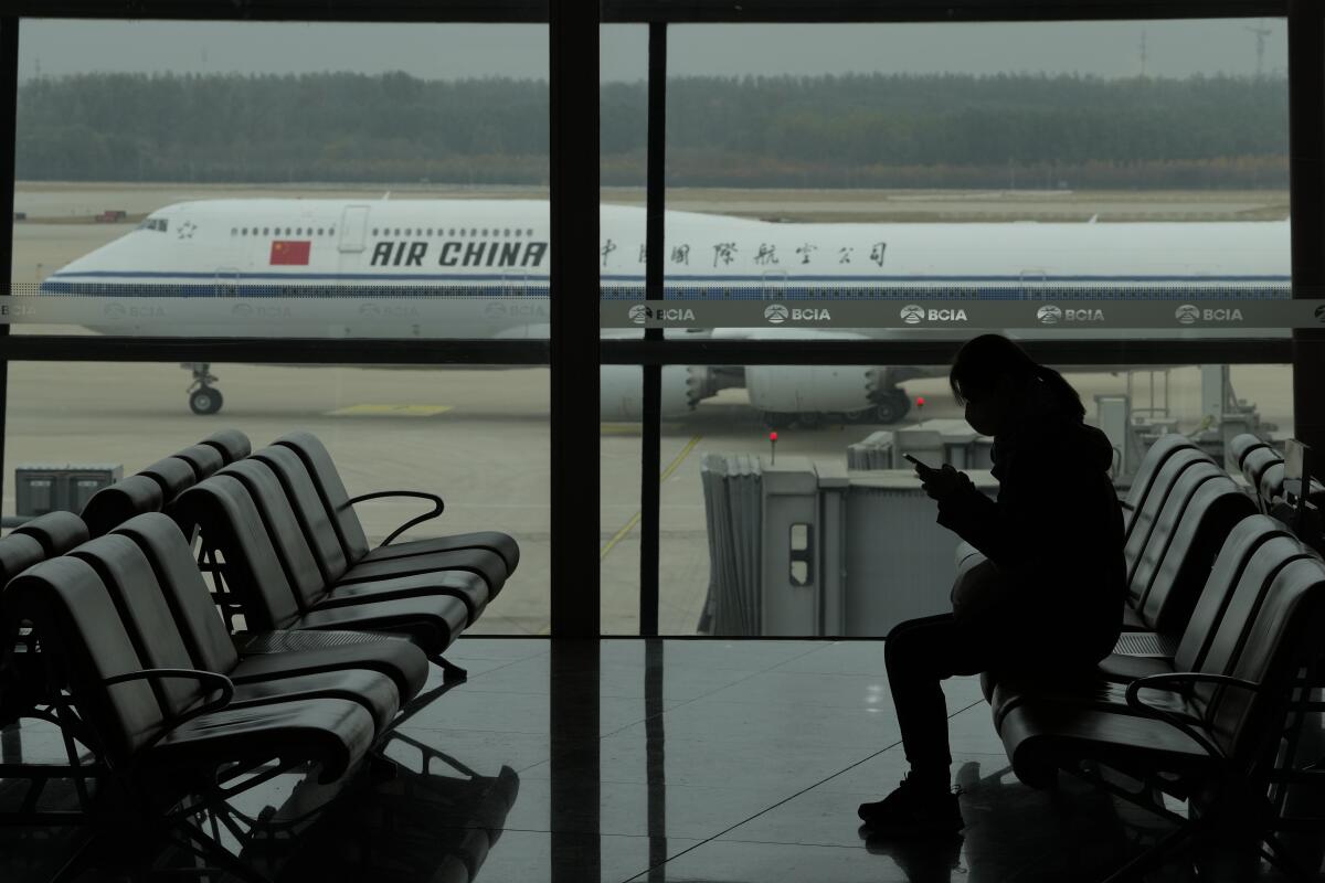 A passenger in a mask sits in an airport as an Air China plane taxis outside.