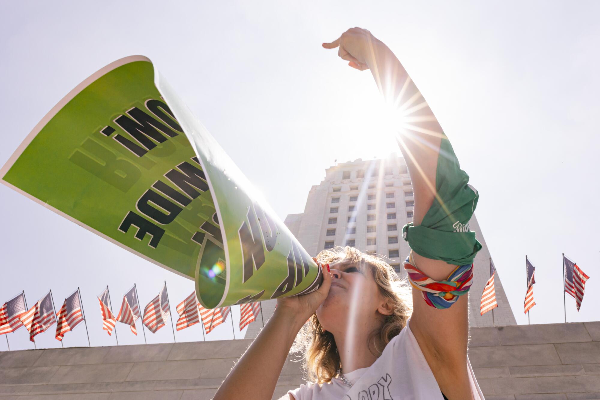 A protester raising her arm and using a poster as a megaphone outside a tall building with a long row of American flags