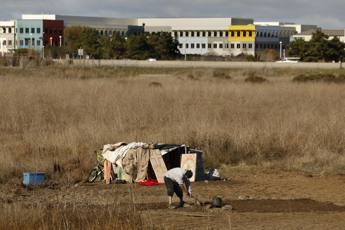 A person next to a makeshift shelter in a field with a building in the background