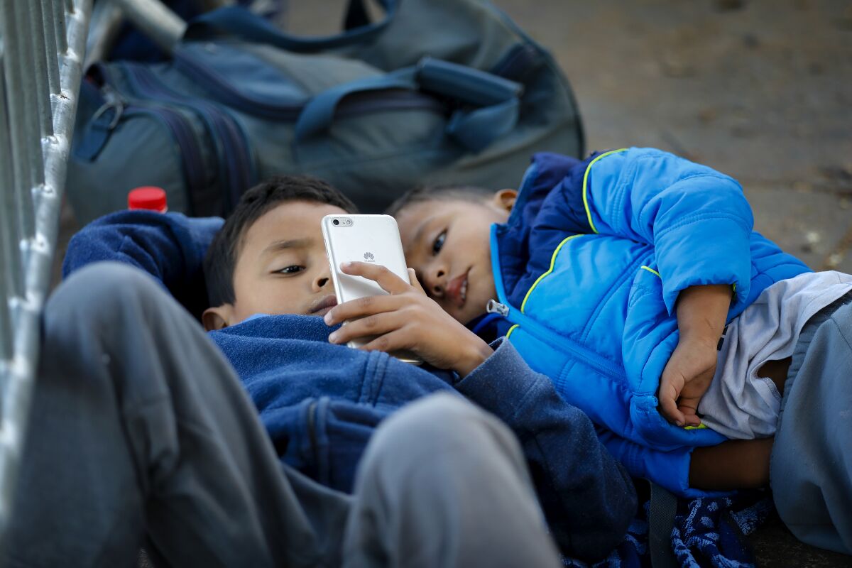 At the El Chaparral port of entry in Mexico on Thursday, October 24, 2019, two boys pass the time by watching a video on a smartphone as they wait with their parents for their number to possibly be called. The boys are part of larger group of migrants that arrive everyday hoping that their number is called to begin the asylum application process to enter into the U.S.