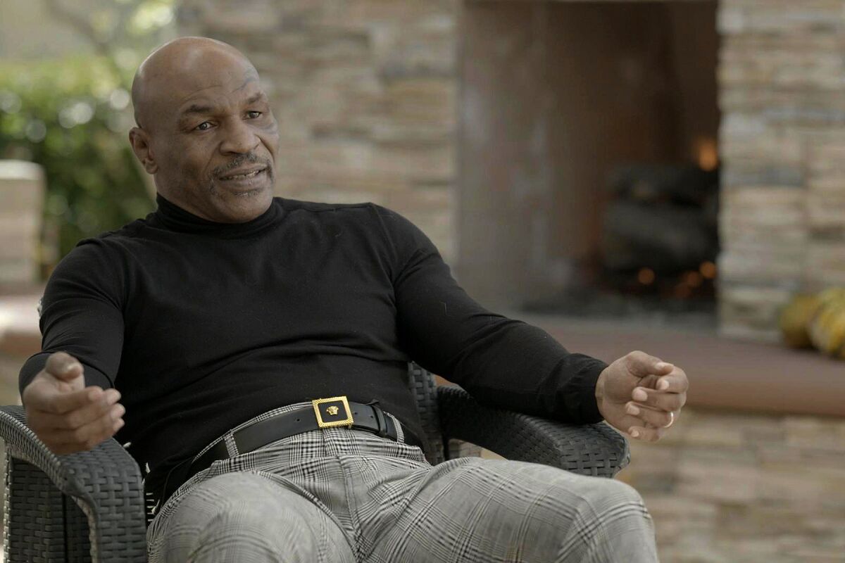 Mike Tyson, wearing a black long-sleeved shirt and plaid pants, sits in a chair.