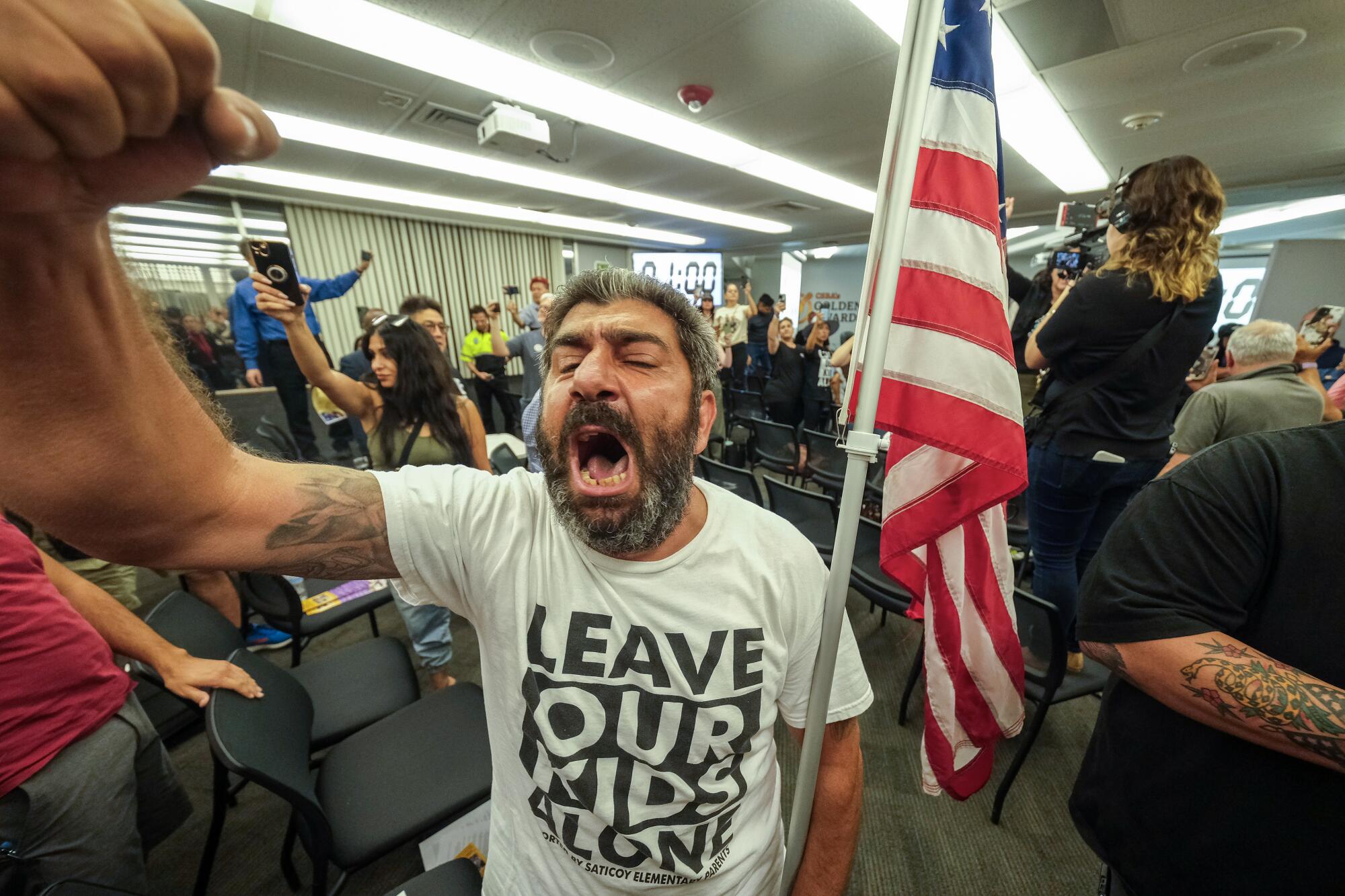 A man wearing a "Leave our kids alone" T-shirt raises a clenched fist and shouts in a crowded meeting room.