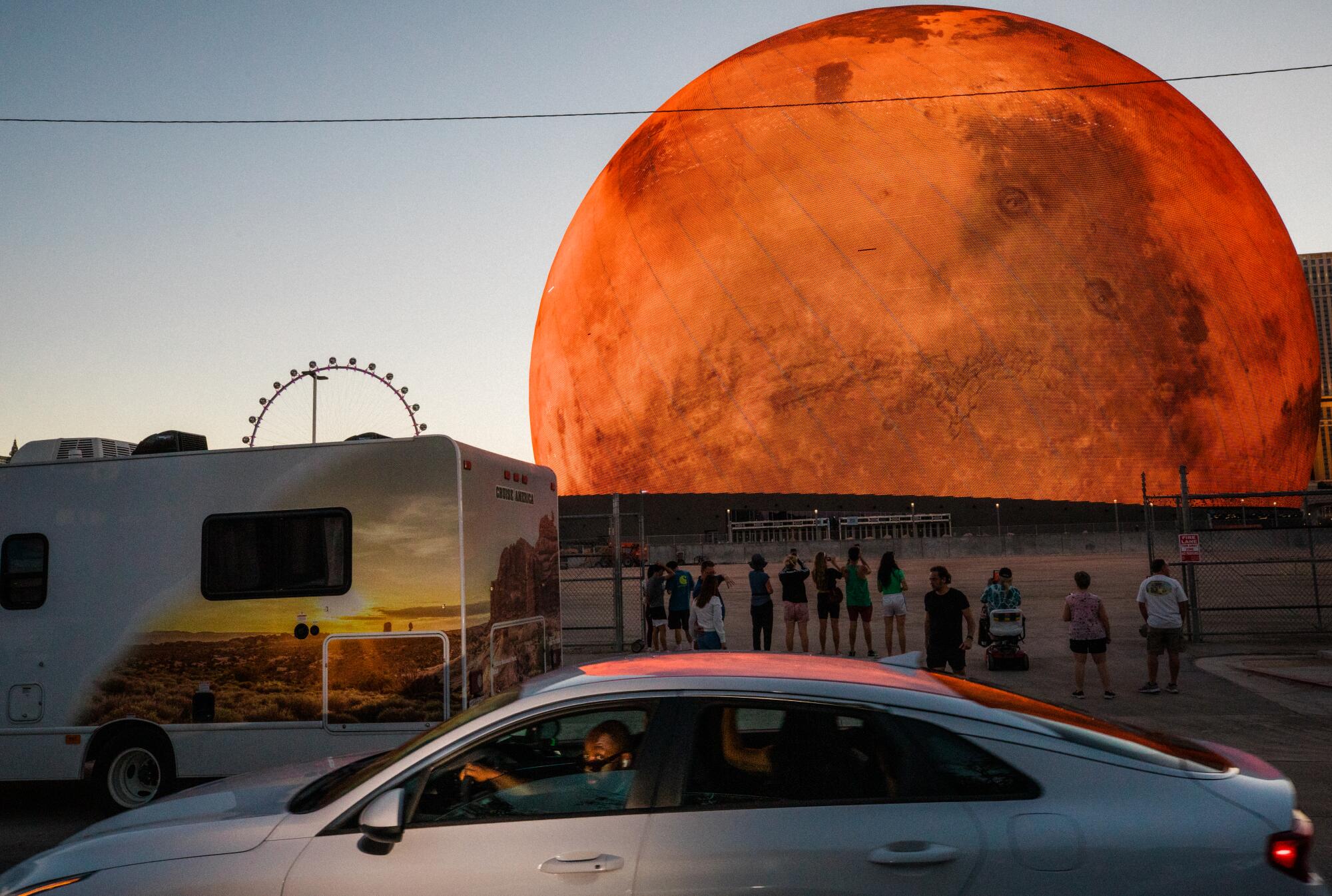 People gather on a street at dusk to watch Sphere broadcast images of a red planet. 
