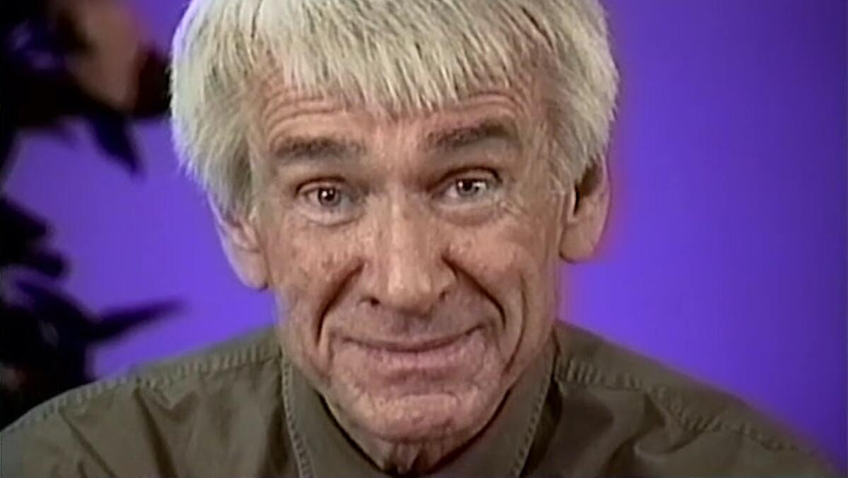 A white-haired man looks directly in a camera before a purple background.