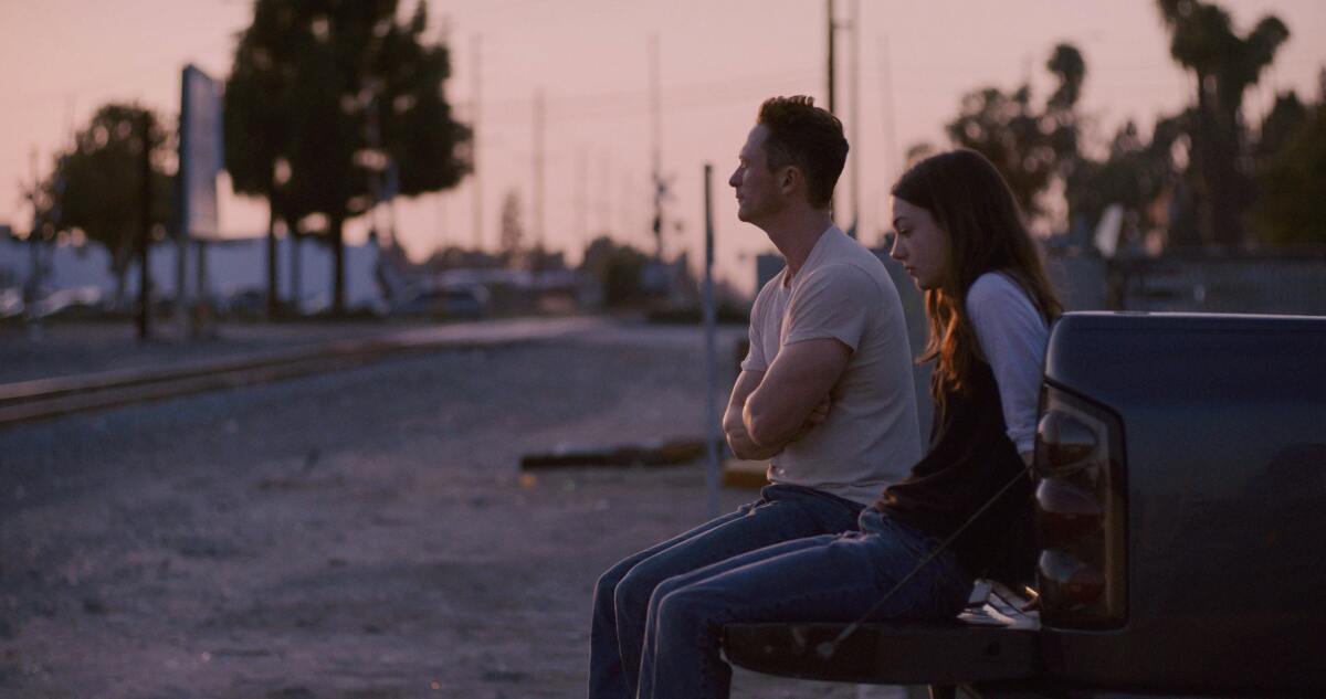 A young man and a girl sit outside on a bench at dusk.