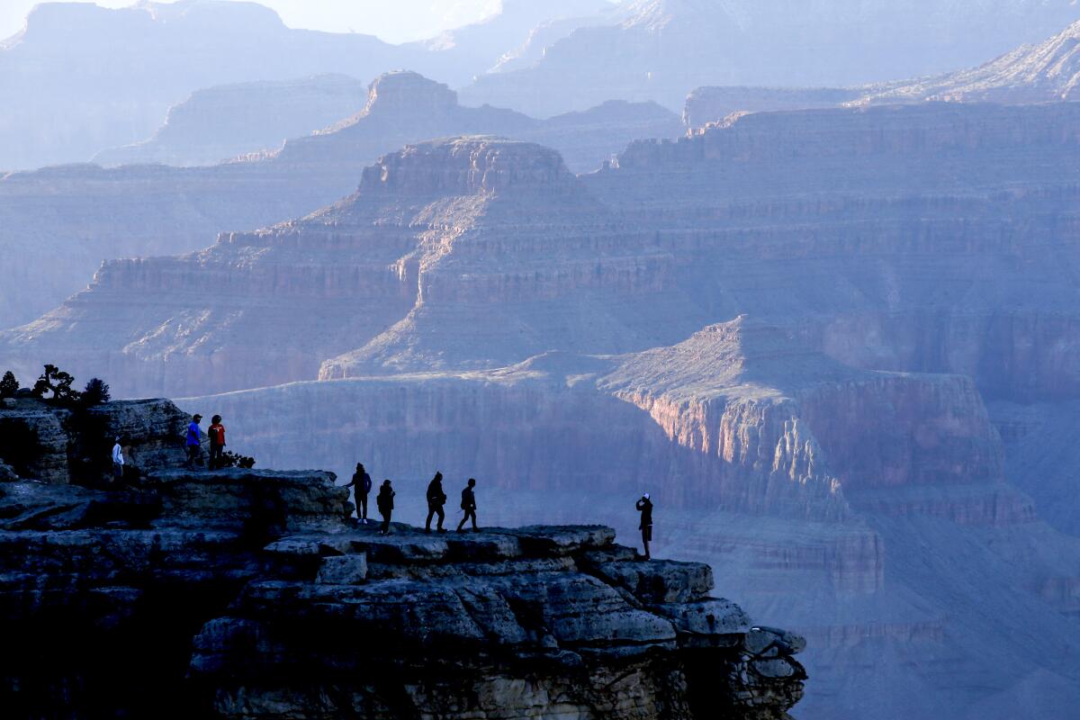 People stand on an outcropping on the rim of the Grand Canyon, seen from a distance