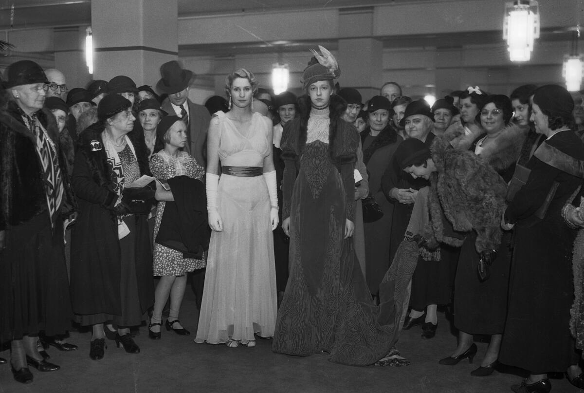 Two women wear long gowns. A crowd of people, most of them in hats and fur-trimmed coats, surround them.