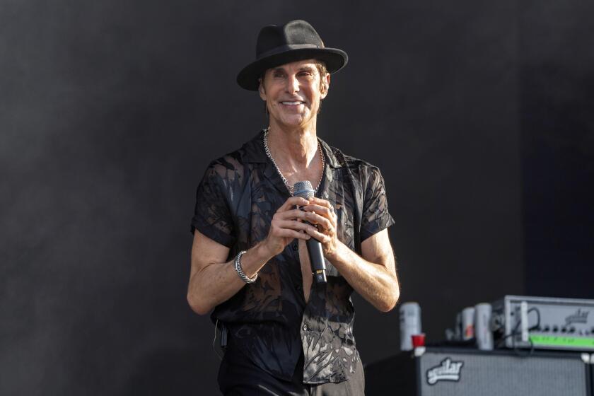 Perry Farrell in a black fedora and a black button-down shirt. He hold a microphone and smiles on a stage