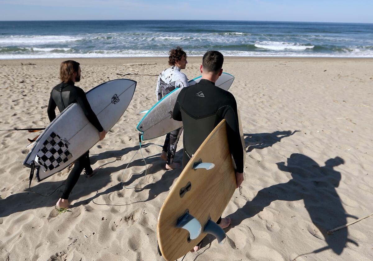 Three men carry surfboards on a beach and walk toward the ocean.