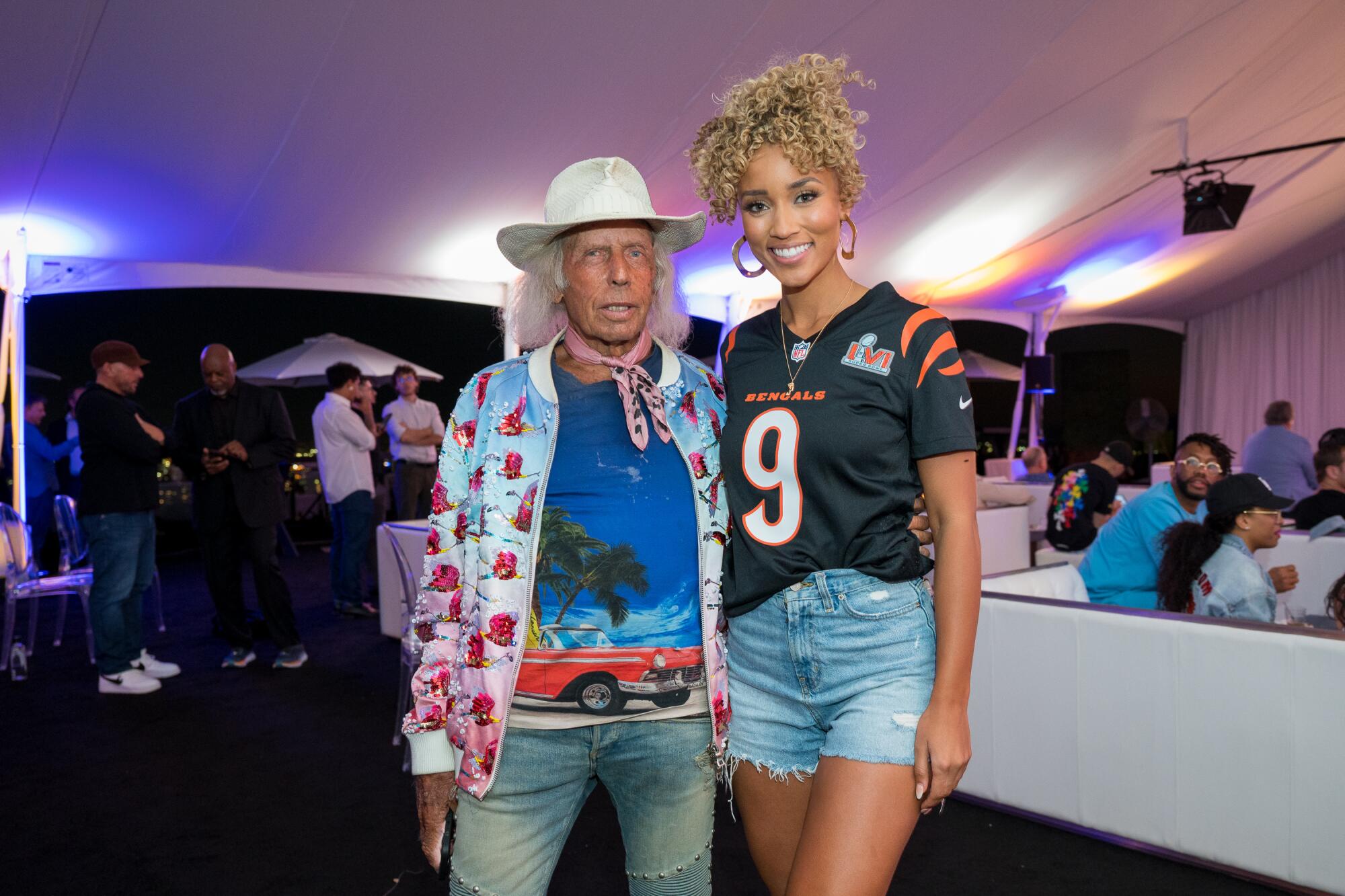 A man in a white hat poses with a woman in a football jersey and cutoff shorts.