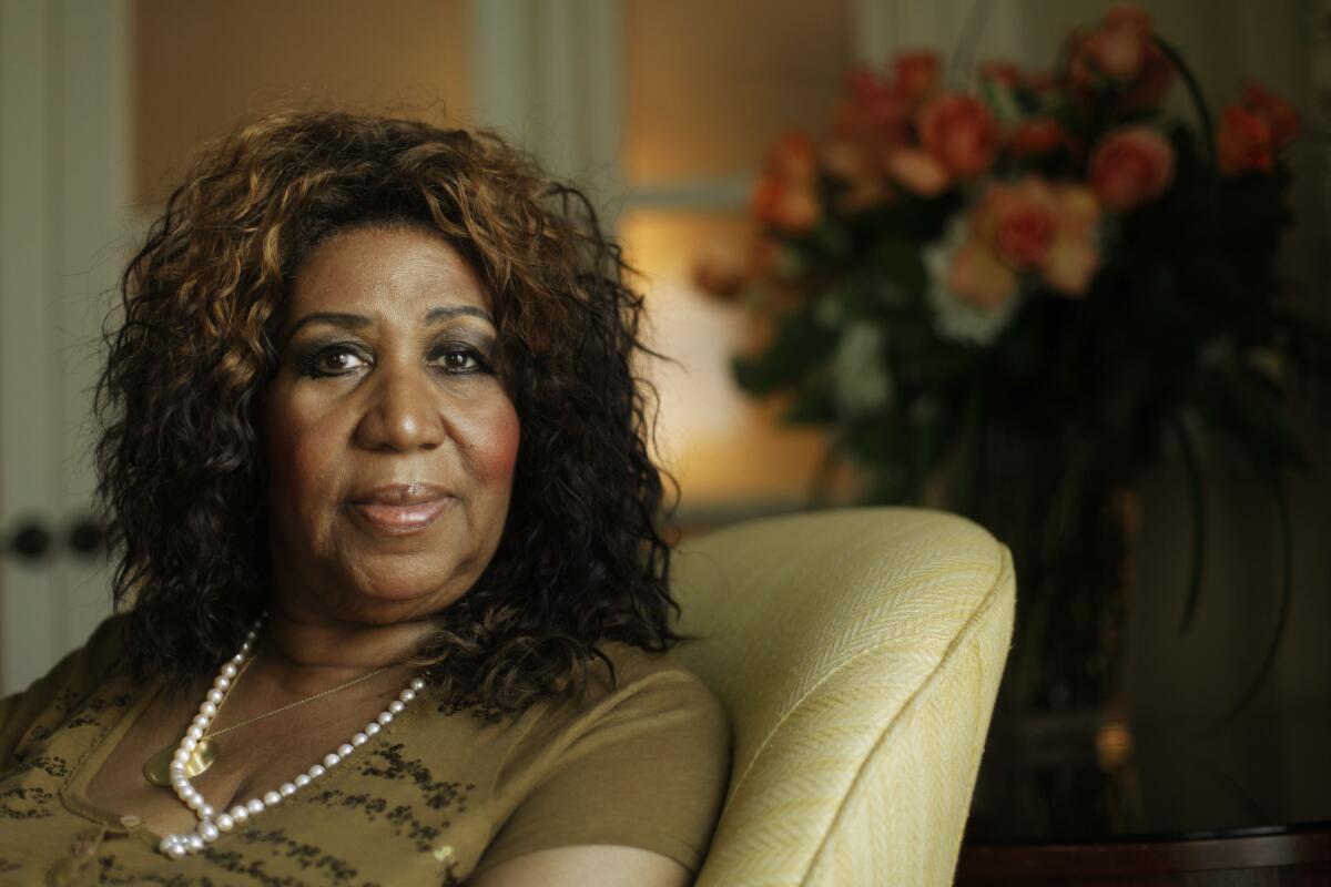 Aretha Franklin looks serious as she sits in a beige chair wearing a brown shirt and pearl necklace