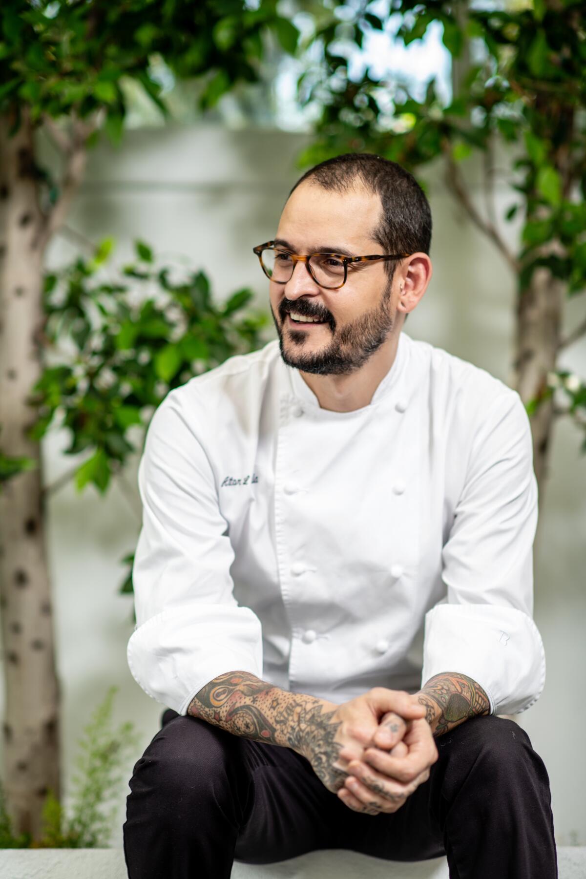 A smiling man in a white chef's coat and black pants looks off camera with his hands clasped.