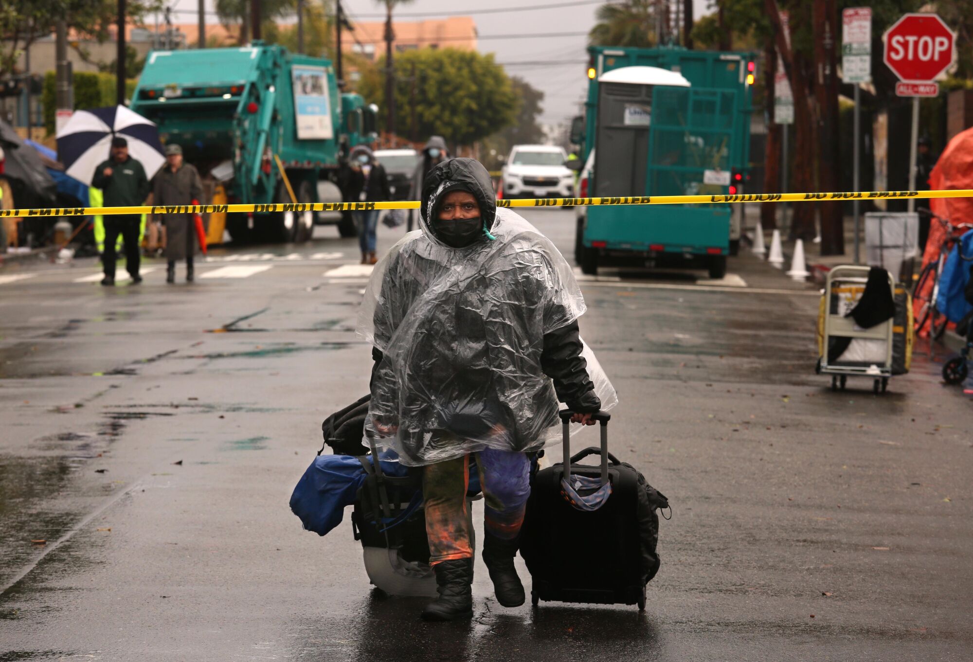 A person in a plastic rain jacket wheels her rolling suitcases. Behind her are green trash trucks and yellow police tape 