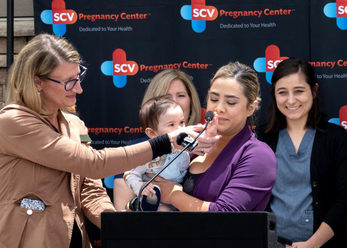 Four women stand behind a portable lectern with a microphone. One is holding a baby who is reaching for the mic.