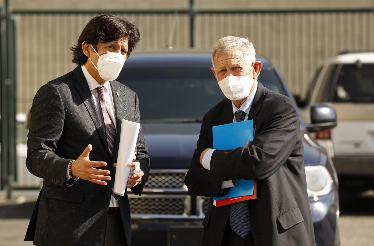 Two men in suits and wearing facemasks stand in a parking lot.