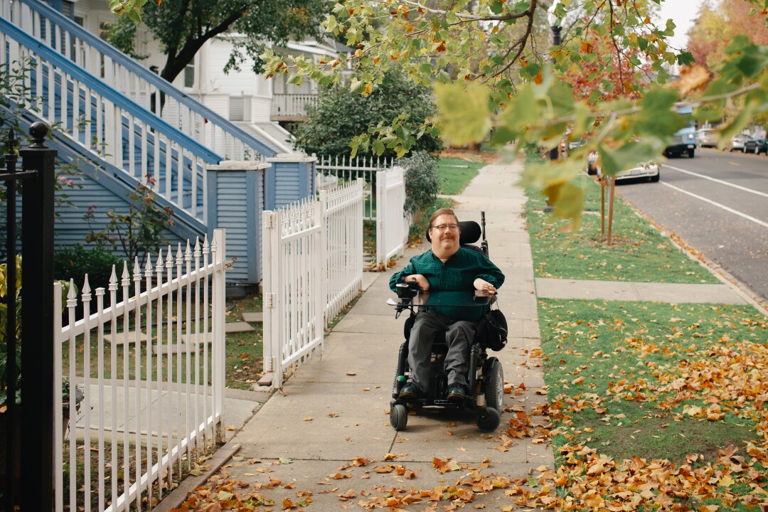 A man sits in a motorized wheelchair on a sidewalk in a residential area