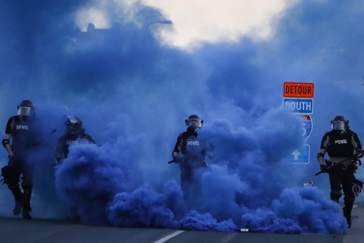 Police in riot gear walk through a cloud of blue smoke