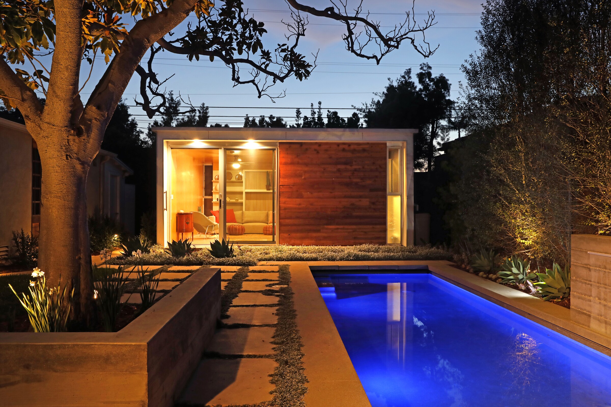 At dusk, a tiny ADU sits in a backyard, just beyond an illuminated lap pool.