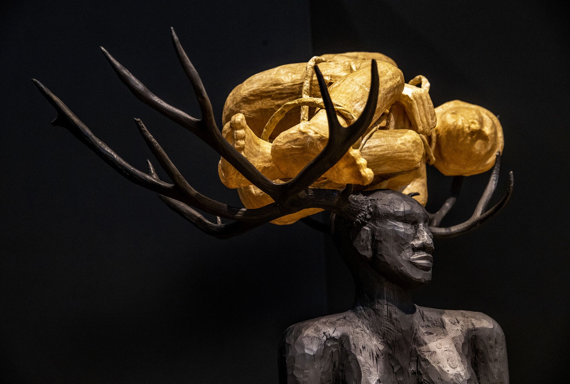 A detail from a wooden sculpture shows a Black woman with antlers supporting the form of another woman on her head