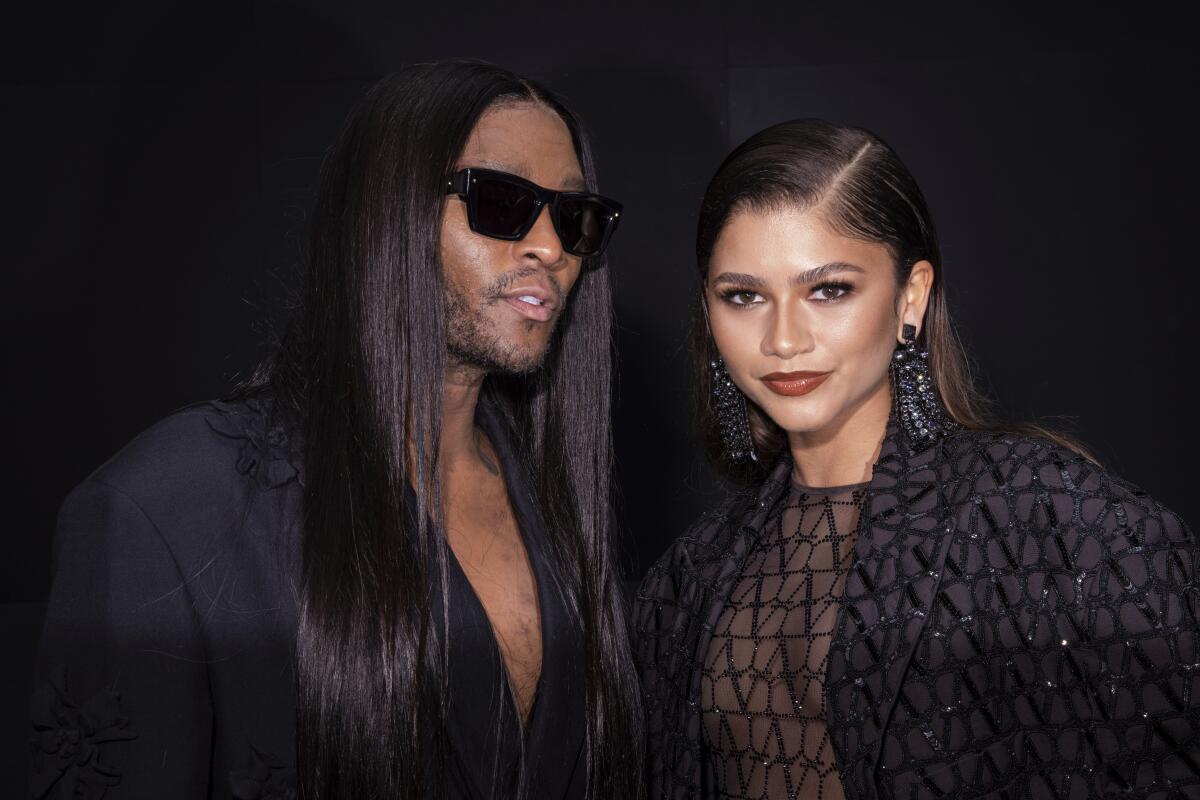 A man with long black hair wearing sunglasses and a suit while posing next to a woman with long brown hair wearing black
