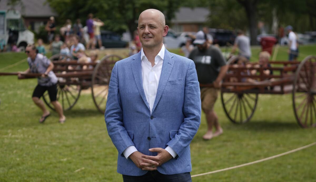 Evan McMullin wears a blue suit on a green field as two people pull wood carts in the background. 