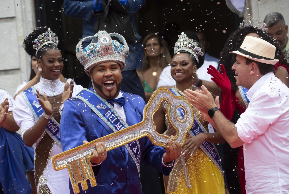 A man wearing a crown and a bright blue suit holds a giant key.