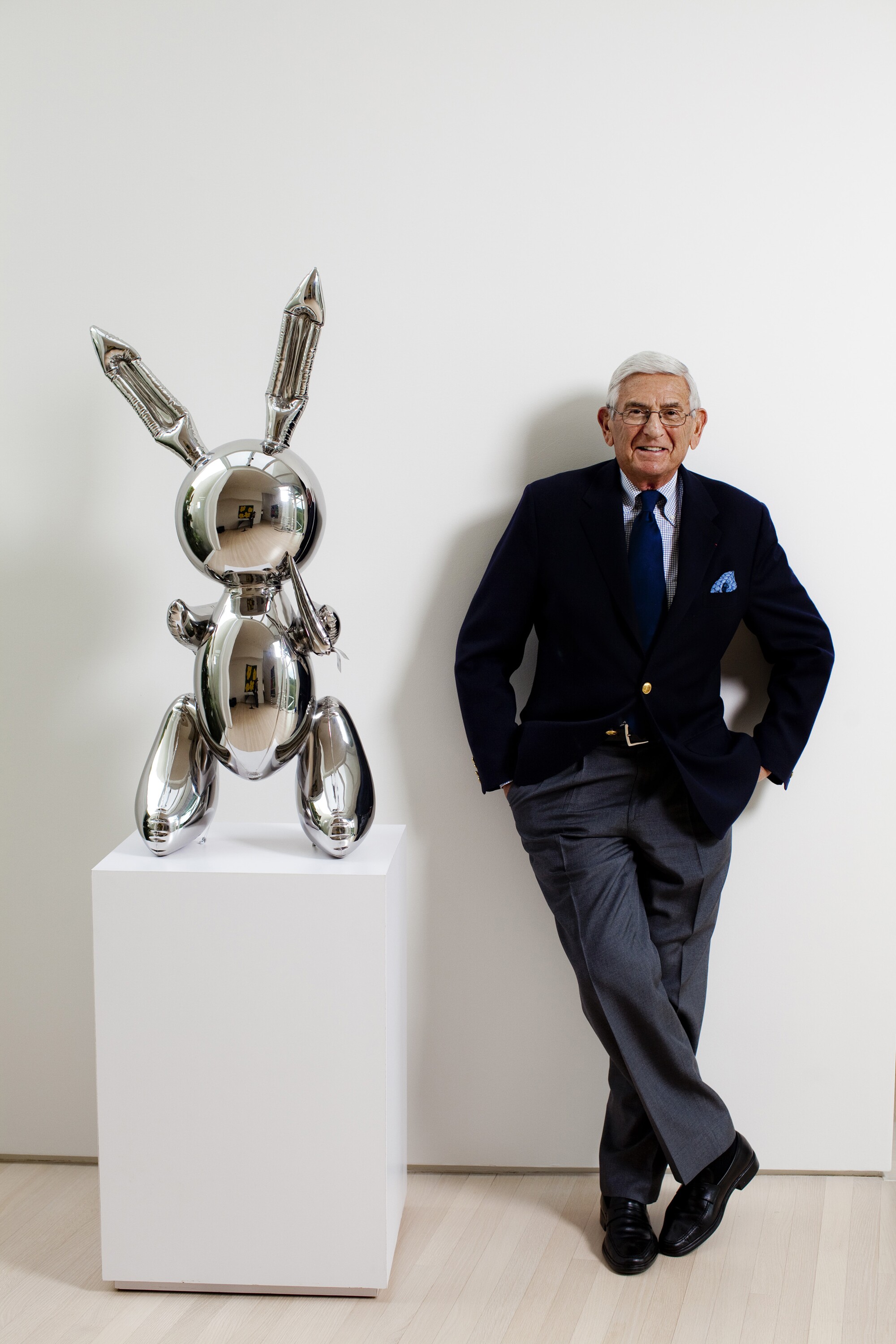 Eli Broad, hands in his pockets, leans against a white wall next to a gray metal sculpture on a plinth.