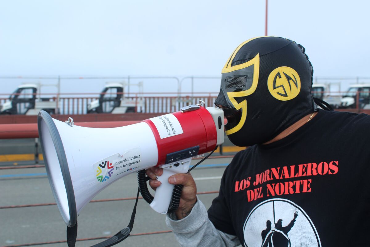 A masked man speaks into a bullhorn while wearing a shirt with Spanish words on it.
