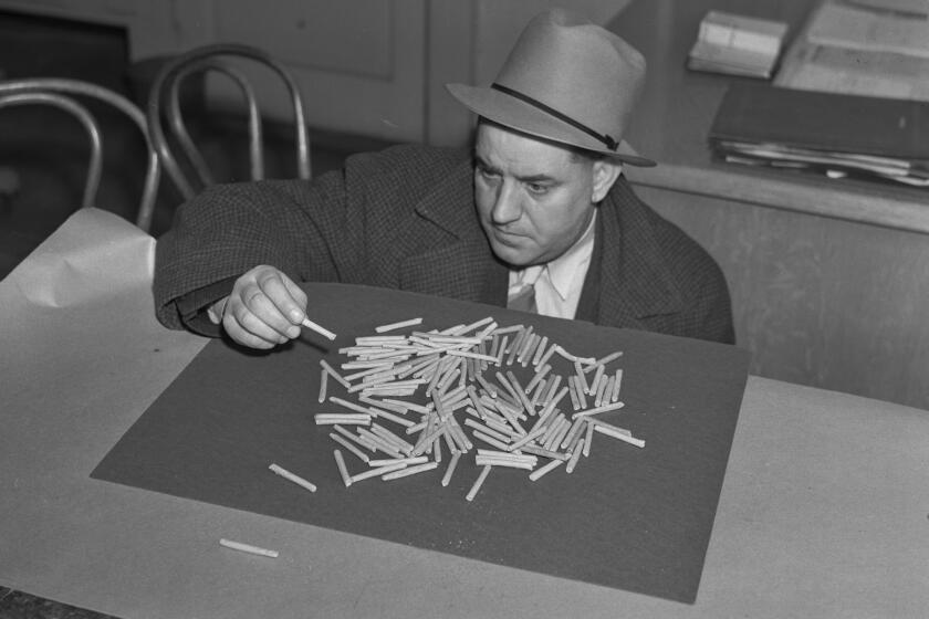 A man in a hat, suit, tie and overcoat leans over a pile of joints on a desktop.