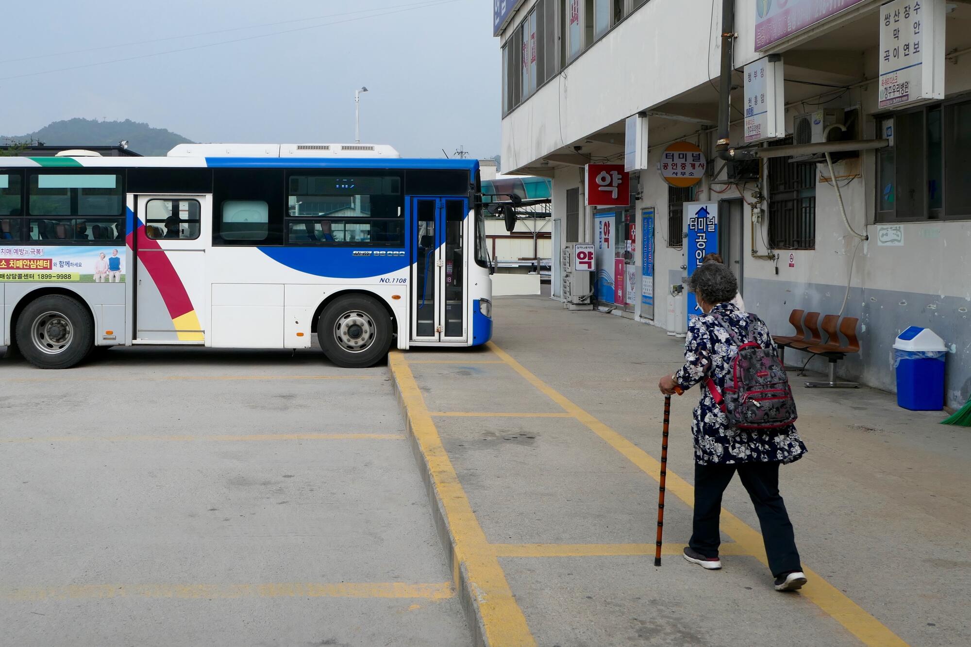 A woman walks with a cane toward a bus next to a building 