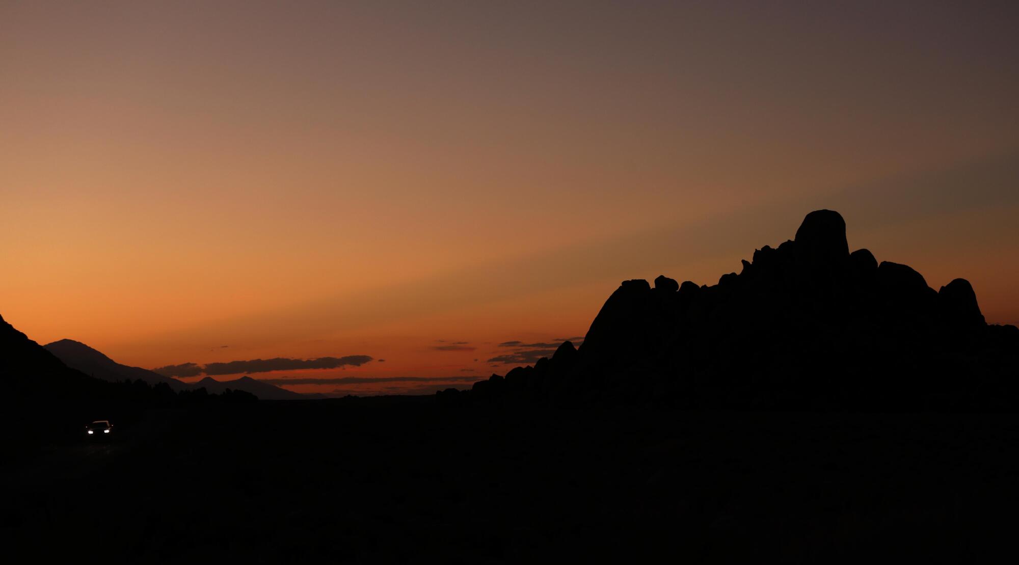 Rock formations are silhouetted against the sunset as a lone car makes its way down a road in the Alabama Hills