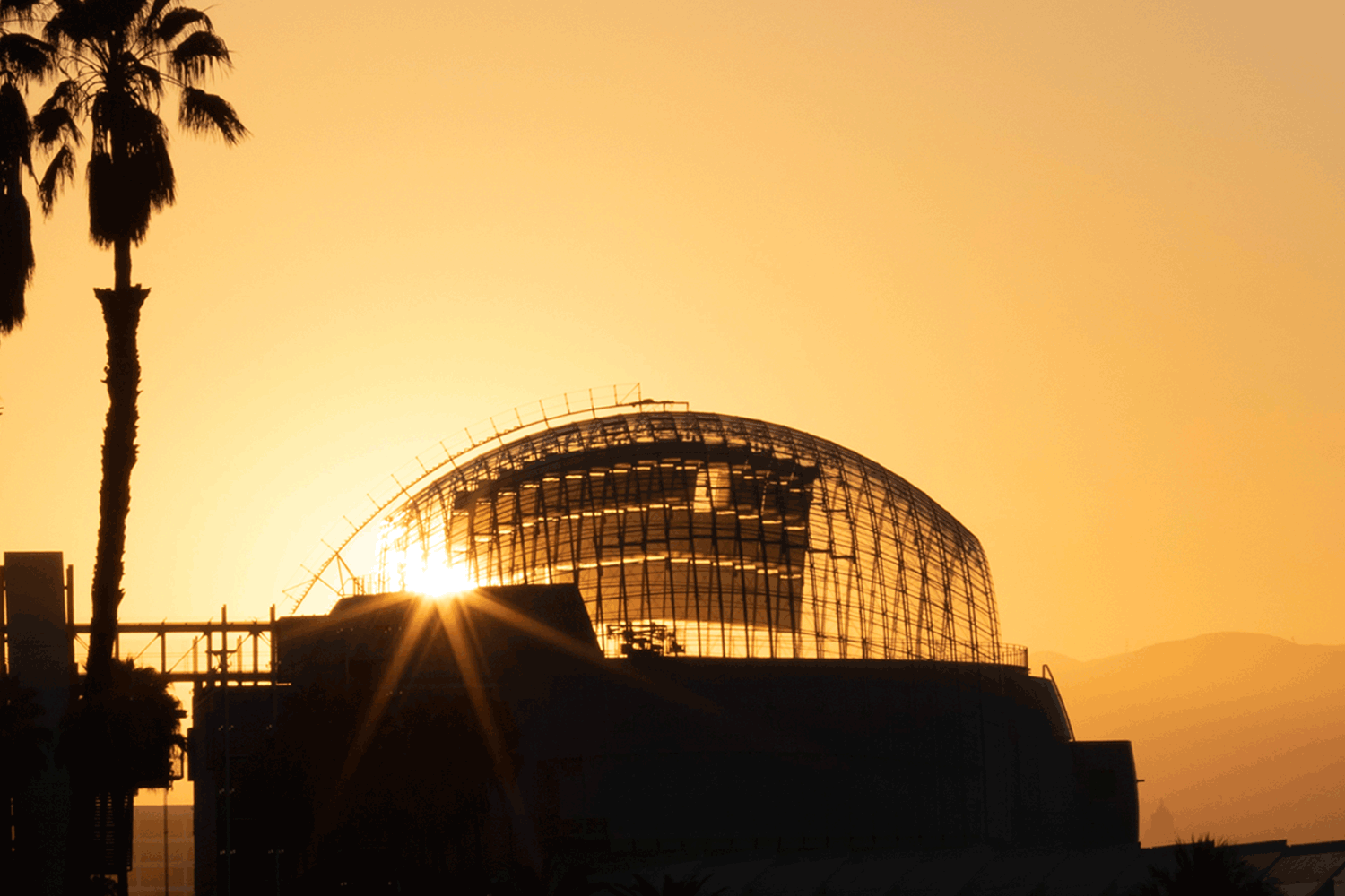 The glass dome of the new Academy Museum terrace appears in silhouette against the setting sun.
