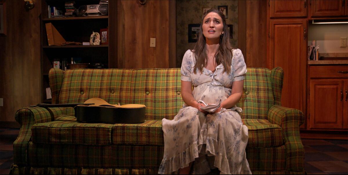 Actor Sara Bareilles, costumed as a pregnant waitress, sits on a couch on a stage set.