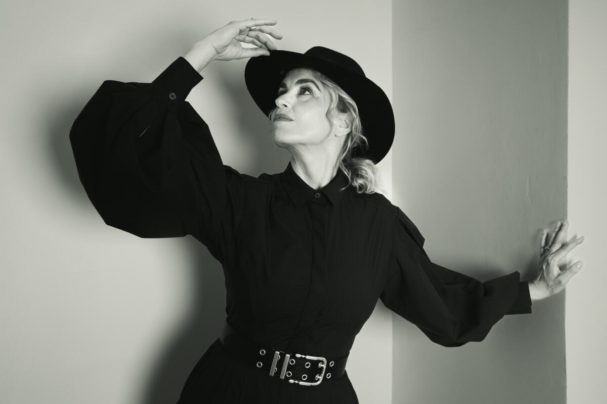 A woman in a black outfit with full sleeves and a belt touches her black hat in a black and white portrait.